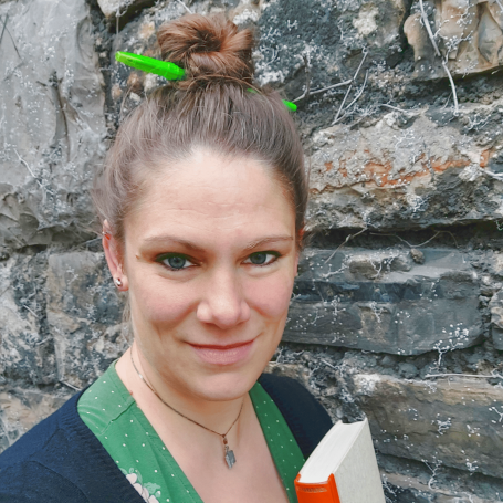 Lektoriel Frau mit hochgestecktem Haar, Halsschmuck, hält ein Buch vor einer Steinfassade.